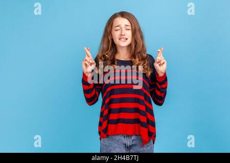 Fortune s'il vous plaît venir.Portrait de la femme en chandail debout avec les doigts croisés pour la bonne chance, faisant le souhait de coeur, priant et espérant pour le miracle.Studio d'intérieur isolé sur fond bleu. Banque D'Images