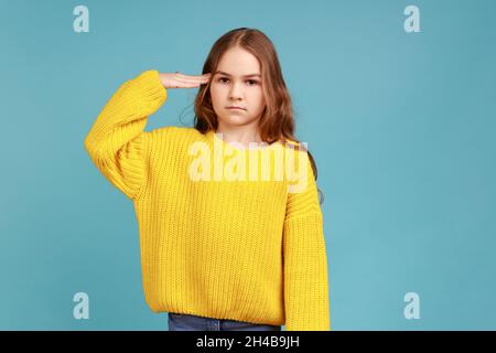 Oui, monsieur.Portrait de petite fille saluant et regardant l'appareil photo avec respect, enfant patriotique, portant jaune décontracté style chandail.Studio d'intérieur isolé sur fond bleu. Banque D'Images