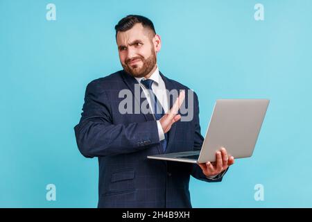 Portrait d'un jeune homme à barbe attrayant portant un costume de style officiel qui s'étire à la main, ne veut pas voir, regarder loin, contenu dégoûtant, studio d'intérieur isolé sur fond bleu. Banque D'Images