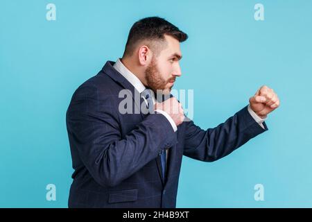 Vue latérale d'un homme d'affaires agressif portant un costume officiel prêt à frapper, à boxe avec des poings serrés, à combattre avec quelqu'un.Studio d'intérieur isolé sur fond bleu. Banque D'Images