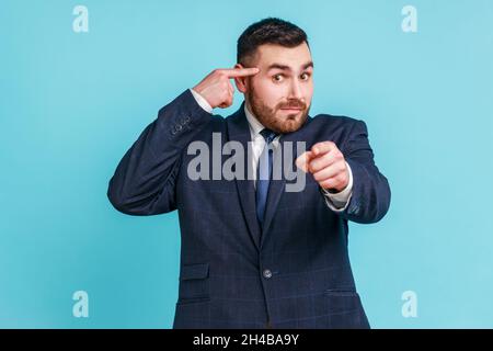 Vous êtes stupide.Triste insatisfait homme d'affaires en costume sombre montrant un geste stupide pointant le doigt vers vous regardant la caméra, blâmant et moquant.Studio d'intérieur isolé sur fond bleu. Banque D'Images