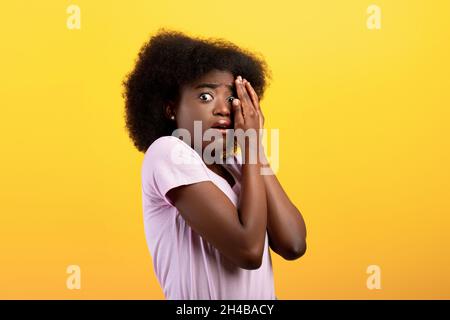 Jeune femme noire stressée couvrant le visage avec les mains et peeking avec un œil, se sentant peur, debout sur fond jaune studio.Femme effrayée tr Banque D'Images