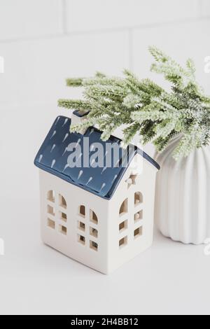 Petite maison de noël en céramique de style scandinave et branches de sapin dans un vase sur une table blanche.Nordic, hygge, maison confortable, concept de confort. Banque D'Images