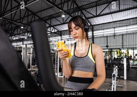 Jeune femme asiatique heureuse assise et écoutant de la musique avec un smartphone tout en faisant de l'exercice à la salle de gym Banque D'Images