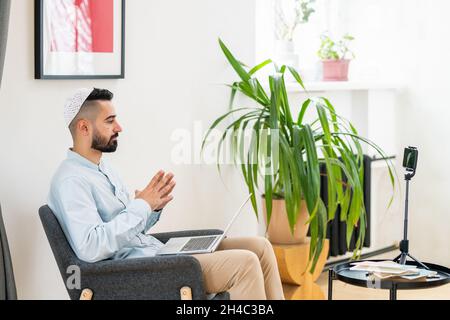 Vue latérale du jeune homme arabe avec un ordinateur portable à genoux assis devant l'appareil photo du smartphone pendant la diffusion en direct Banque D'Images