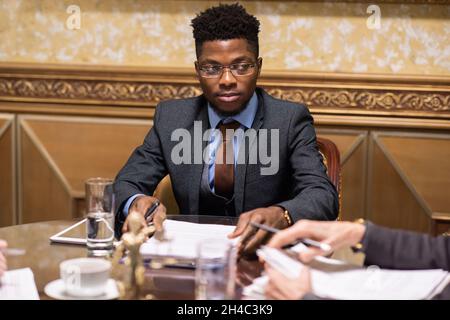 Jeune homme d'affaires sérieux en lunettes et costume élégant regardant un collègue à la réunion tout en étant assis à table Banque D'Images