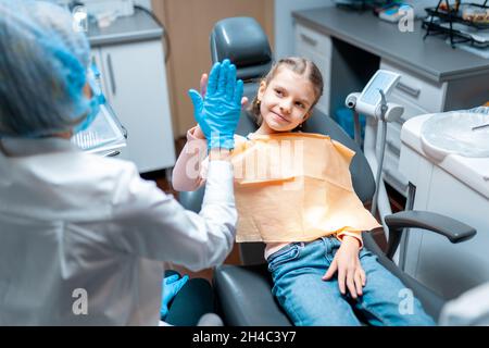 Dentiste amical donnant haute cinq petite fille assise sur une chaise stomatologique après le traitement Banque D'Images