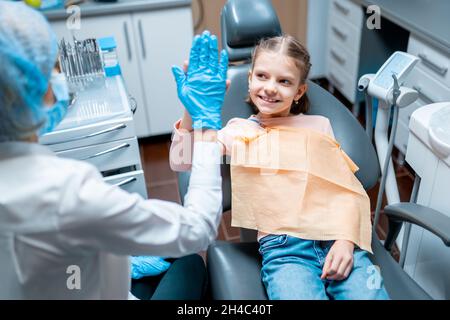 Femme sympathique dentiste donnant élevé cinq petite fille assise sur une chaise stomatologique après le traitement Banque D'Images