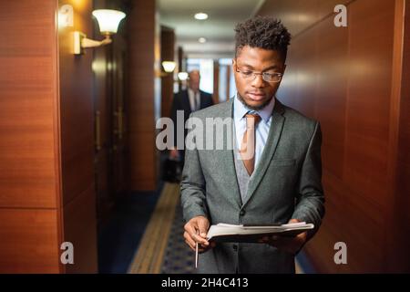 Jeune homme d'affaires africain élégant en vêtements de formalwear et lunettes regardant à travers le document dans le couloir du centre de bureau moderne Banque D'Images