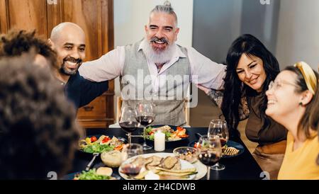 Des amis gais adultes se réjoument de s'asseoir à la table pendant un dîner.Mise au point sélective sur l'homme barbu au centre - image ISO élevée. Banque D'Images