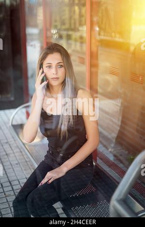 Une jeune fille blonde parle au téléphone en attendant d'être assise à un arrêt d'autobus Banque D'Images