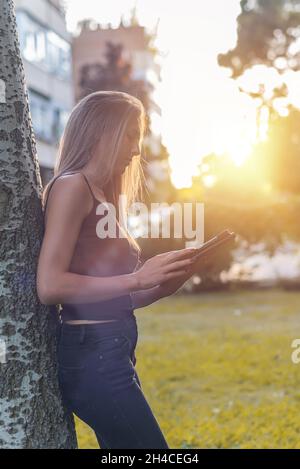 Jolie jeune femme blonde utilisant une tablette au coucher du soleil dans un parc Banque D'Images