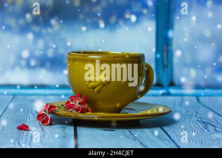 Une tasse jaune, soucoupe se tient sur une table en bois d'épicéa contre une fenêtre.Fond d'écran bleu.Chutes de flocons de neige en soirée d'hiver.Petite fleur P Banque D'Images