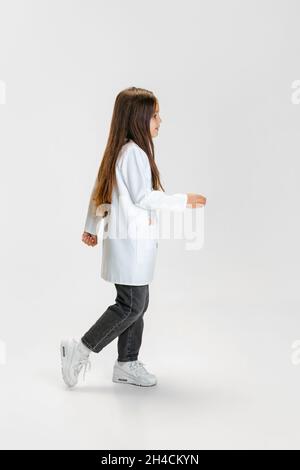 Vue latérale.Mignon belle petite fille, enfant à l'image d'un médecin portant un manteau blanc de laboratoire posant isolé sur fond blanc de studio Banque D'Images