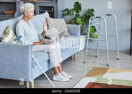 Vue latérale portrait en longueur d'une femme âgée aux cheveux blancs utilisant un ordinateur portable à la maison et en agitant à l'appareil photo, espace de copie Banque D'Images