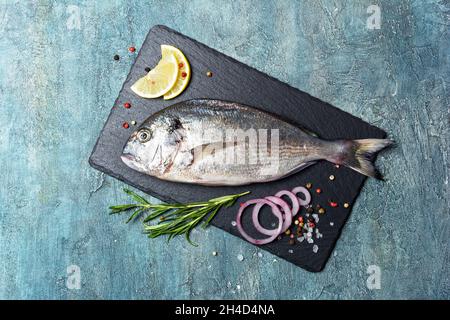 Vue de dessus de poisson frais dorado ou dorade à tête dorée avec citron, oignon et épices comme poivre, romarin, sel sur ardoise noire et fond de béton bleu Banque D'Images