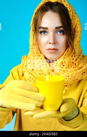 femme dans un foulard et un chandail jaune tient une tasse dans ses mains. fond bleu Banque D'Images
