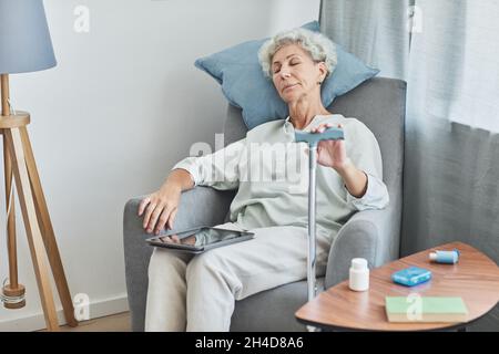 Portrait d'une femme âgée aux cheveux blancs dormant dans un fauteuil confortable à la maison avec canne, espace de copie Banque D'Images