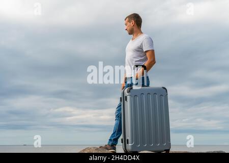Un jeune homme en Jean et une chemise blanche se tient avec une valise grise sur le bord de mer sur d'énormes rochers contre un ciel avec des nuages et regarde dans la distance Banque D'Images