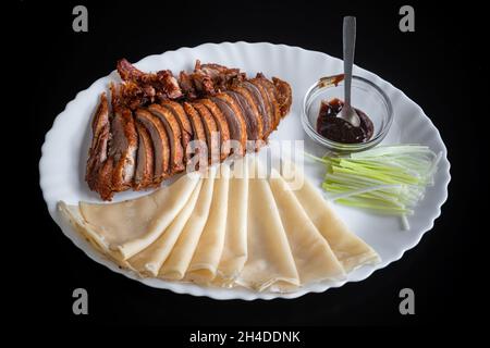 Canard de Pékin, canard croustillant frit à la chinoise, servi avec une sauce hoisine, des crêpes et du concombre, dans une assiette blanche sur fond noir. Banque D'Images