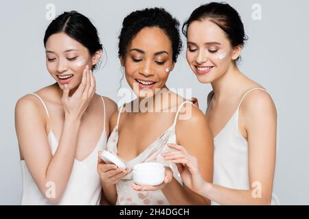 Joyeuses femmes multiculturelles appliquant de la crème cosmétique sur les visages isolés sur le gris Banque D'Images