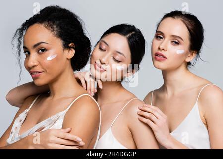 Femme afro-américaine souriante debout près d'amis interraciaux avec de la crème sur des visages isolés sur le gris Banque D'Images