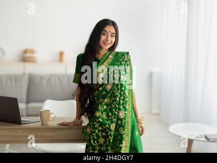 Portrait de la belle dame indienne en robe verte sari s'appuyant sur la table et souriant à l'appareil photo, à l'intérieur Banque D'Images