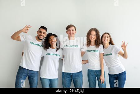 Groupe de jeunes heureux divers bénévoles embrassant et posant sur le mur du studio léger, montrant des gestes ok Banque D'Images