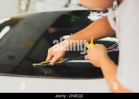 Un spécialiste masculin applique un film de coloration de fenêtre sur la voiture et coupe l'excédent de pièce à l'aide d'un couteau. Banque D'Images