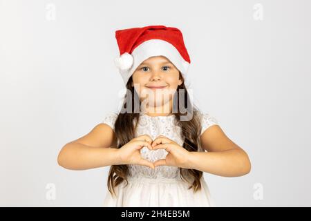 Portrait de fille heureuse et douce en robe blanche, portant le chapeau rouge du Père Noël montre le signe du coeur des doigts isolés sur fond blanc avec un endroit vide Banque D'Images