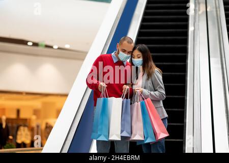 Jeune couple international dans des masques de protection descendant sur l'escalier roulant au centre commercial, regardant à l'intérieur des sacs de shopping, espace libre Banque D'Images