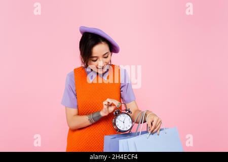 femme tendance en robe sans manches orange mettant un réveil vintage dans un sac de shopping isolé sur rose Banque D'Images