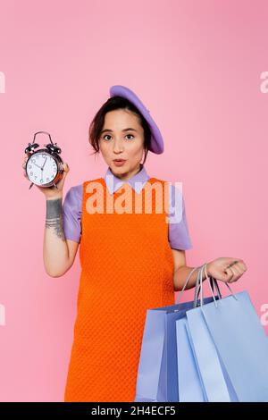 femme stupéfait avec des sacs de shopping montrant le réveil vintage isolé sur rose Banque D'Images