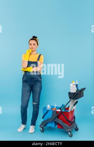 nettoyer les gants en caoutchouc et les combinaisons en denim en regardant l'appareil photo près du chariot avec les fournitures de nettoyage en bleu Banque D'Images