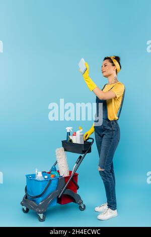 nettoyant dans les combinaisons en denim et les gants en caoutchouc en portant le selfie près du chariot avec les fournitures de nettoyage en bleu Banque D'Images