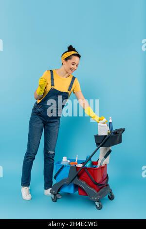 nettoyant pour jeunes dans des combinaisons en denim souriant près du chariot avec fournitures de nettoyage en bleu Banque D'Images