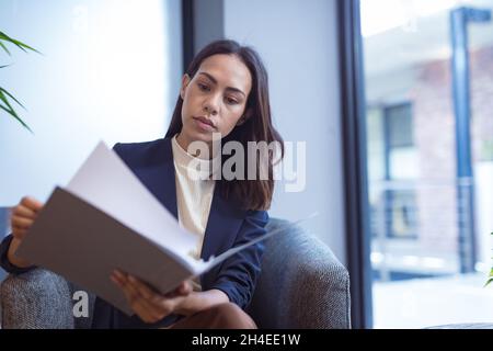 Femme d'affaires biraciale vérifiant des documents avec des collègues en arrière-plan dans un bureau moderne Banque D'Images