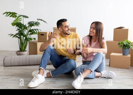 Joyeux jeune couple divers assis dans le plancher avec des verres de champagne, toaster, célébrant la réinstallation dans la nouvelle maison Banque D'Images