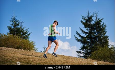 Coureur de sentier qui s'aventure dans la prairie en plein air sous le ciel bleu et regarde sa montre.Jogging dans un espace nature ouvert au soleil par temps chaud.Concept de sport, d'entraînement et de loisirs actifs. Banque D'Images