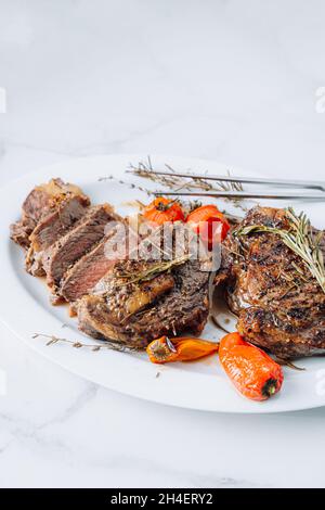 steaks de ribeye juteux bien frits sur un plateau blanc sur une surface en marbre blanc avec tomates cerises et poivrons raspasmarins et frits. les steaks sont tranchés Banque D'Images