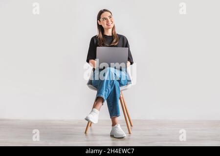 Belle jeune femme assise sur une chaise, travaillant sur un ordinateur portable contre un mur blanc de studio, pleine longueur Banque D'Images
