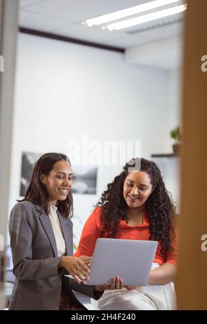 Deux femmes d'affaires souriantes et diverses qui s'assoyent et utilisent un ordinateur portable dans un bureau moderne Banque D'Images