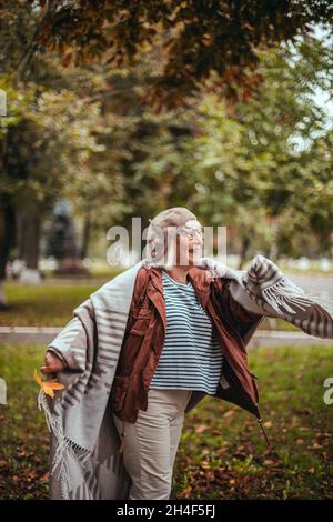 Saison d'automne.Femme adulte élégante et heureuse des années 50 dans un béret en laine et des vêtements de boho confortables marchant dans le parc de la ville d'automne.Accessoires pour lunettes Lady in minimaliste Banque D'Images