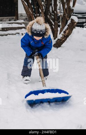 Petit garçon avec une pelle à la main enlever la neige dans l'arrière-cour, déneigement.Enfant en blouson bleu nettoie la neige avec une pelle après une chute de neige Banque D'Images