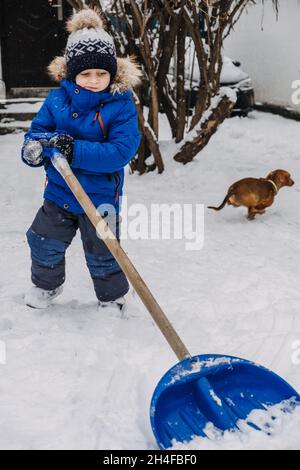 Petit garçon avec une pelle à la main enlever la neige dans l'arrière-cour, déneigement.Enfant en blouson bleu nettoie la neige avec une pelle après une chute de neige Banque D'Images