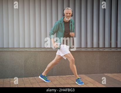 Prise de vue en longueur d'un homme mûr et gai dans des vêtements de sport et des écouteurs se prépare à l'entraînement en plein air Banque D'Images