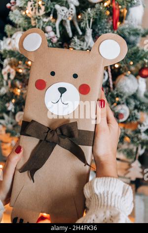 Photo perspective personnelle de la femme tenant le cadeau de Noël enveloppé comme un ours devant un arbre de noël décoré. Banque D'Images