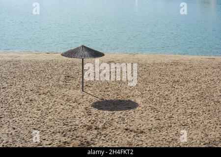 Un parasol en roseau sur une plage vide à midi sous le soleil d'automne, jetant une grande ombre.Solitude. Banque D'Images