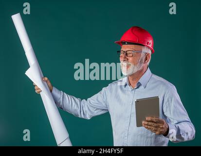 Portrait d'un ingénieur senior portant un casque et tenant des bleus en rouleau et en tablette sur fond bleu Banque D'Images