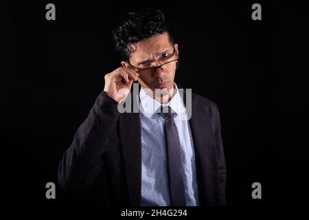 Homme d'affaires prenant des lunettes et regardant de manière suspecte, isolé sur fond noir.Un jeune latins adulte fronce des sourcils avec un regard distrayant Banque D'Images